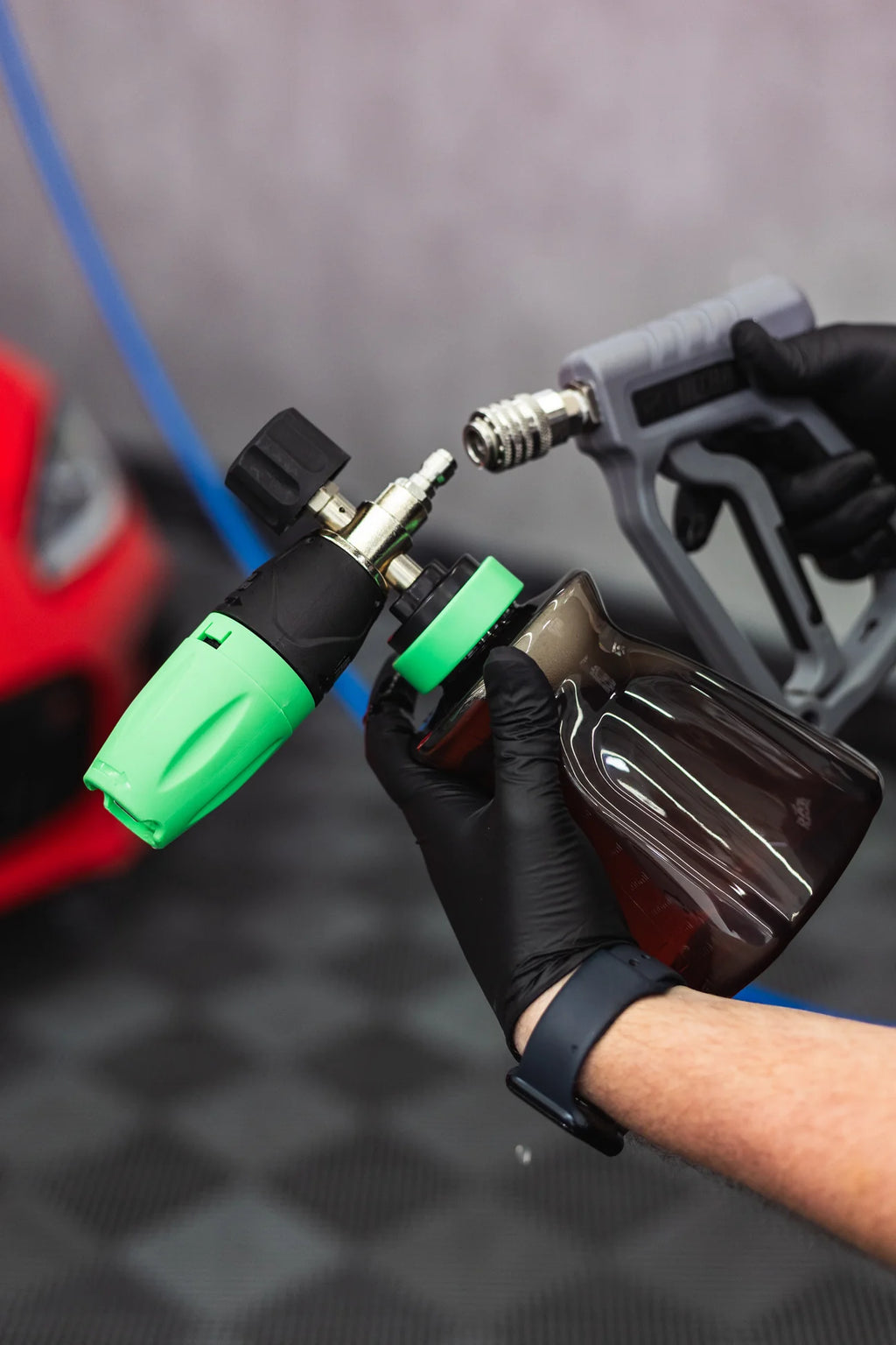 Gloved hand attaching a green and black foam cannon with a brown bottle to a pressure washer spray gun.