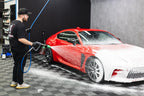 Detailer using a foam cannon pressure washer to cover a red sports car in thick white cleaning foam inside a detailing bay.