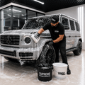 A man in a black t-shirt and cap washes a silver Mercedes-Benz G-Class SUV covered in soap foam inside a modern garage. Two buckets in the foreground are labeled 'SUPREME DETAILING SUPERSTONE' and 'RINSE'.